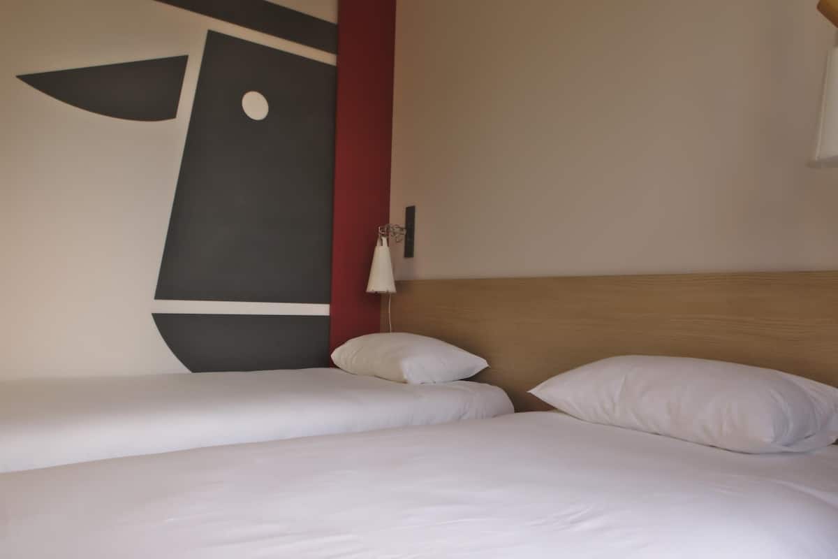 Een hotelkamer met twee enkele bedden, een houten hoofdeinde, een wandlamp en een groot abstract kunstwerk.