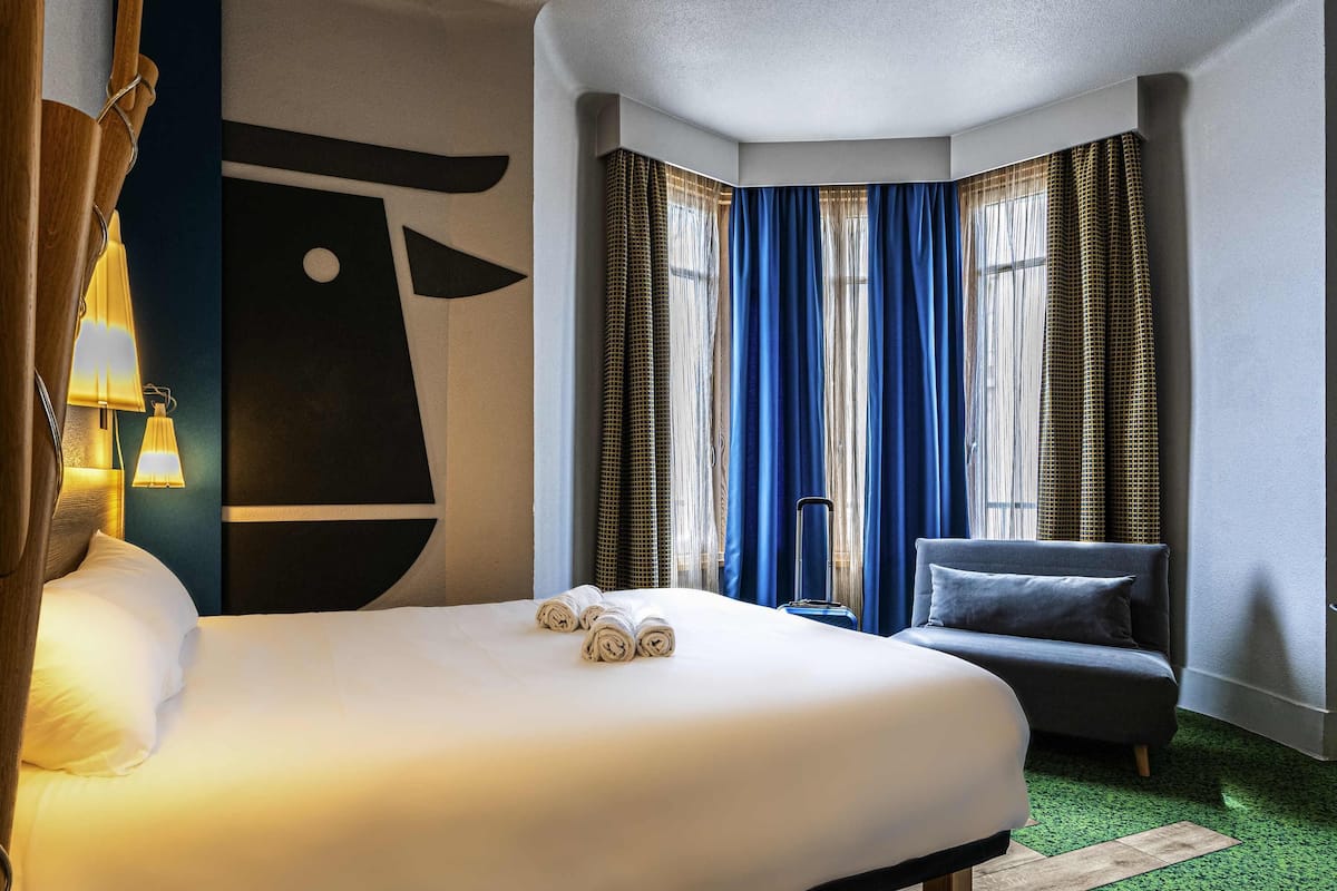 Een hotelkamer met een groot bed, een kleine bank, een koffer en een raam met blauwe gordijnen.
