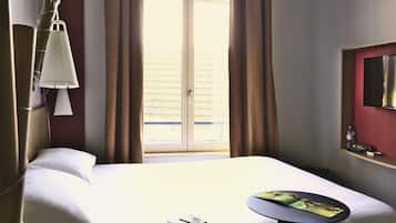 Habitación de hotel con cama, mesita de noche con teléfono y revista, lámpara, ventana con cortinas y televisor en la pared.