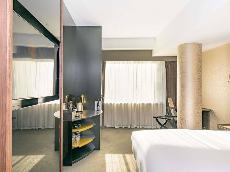 A hotel room with a bed, a desk, a mirror, a minibar, and a large window with curtains.