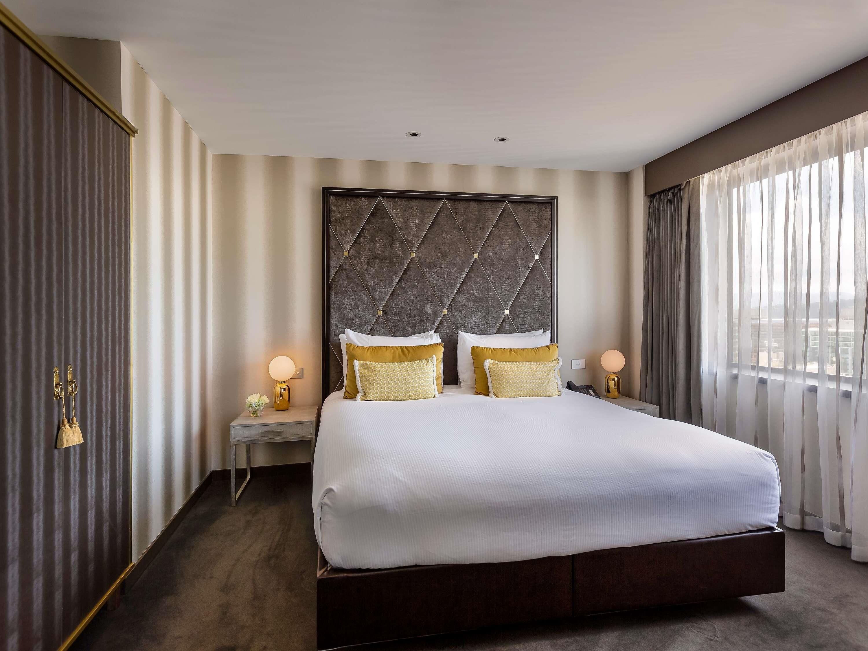 A hotel room with a large bed, a headboard with a geometric pattern, bedside lamps, and sheer curtains.