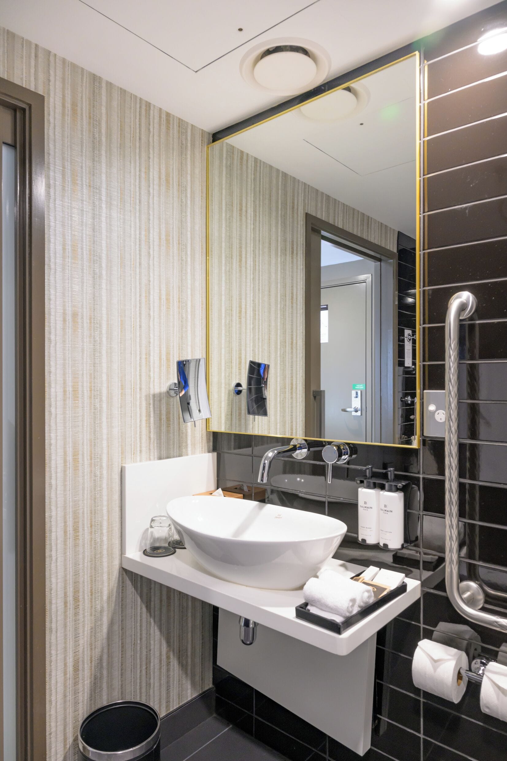 A modern bathroom with a large white sink, a mirror with a gold frame, a towel rack, and a toilet paper holder.