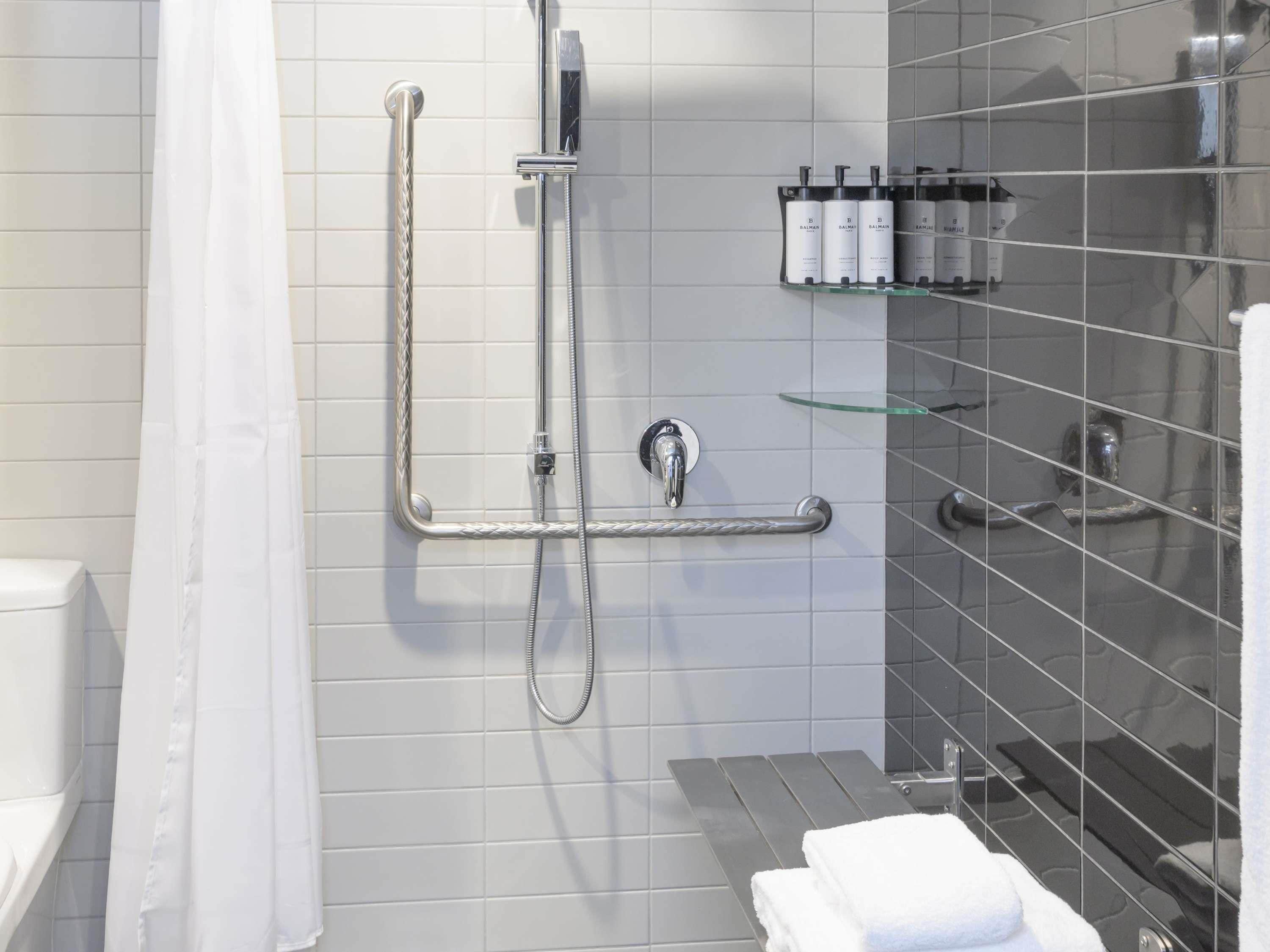 A modern bathroom with a walk-in shower, grab bars, a glass shelf with toiletries, and a white towel on a metal rack.