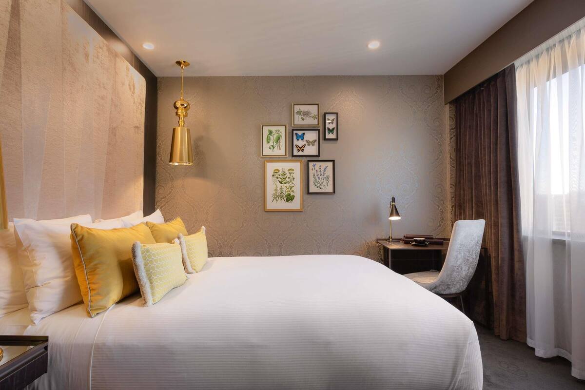A hotel room with a large bed, a desk, a chair, and framed artwork on the wall.
