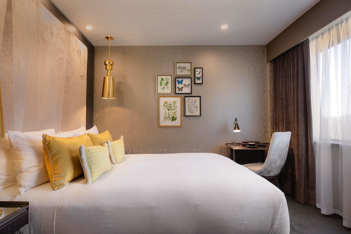 A hotel room with a large bed, a desk, a chair, and framed artwork on the wall.