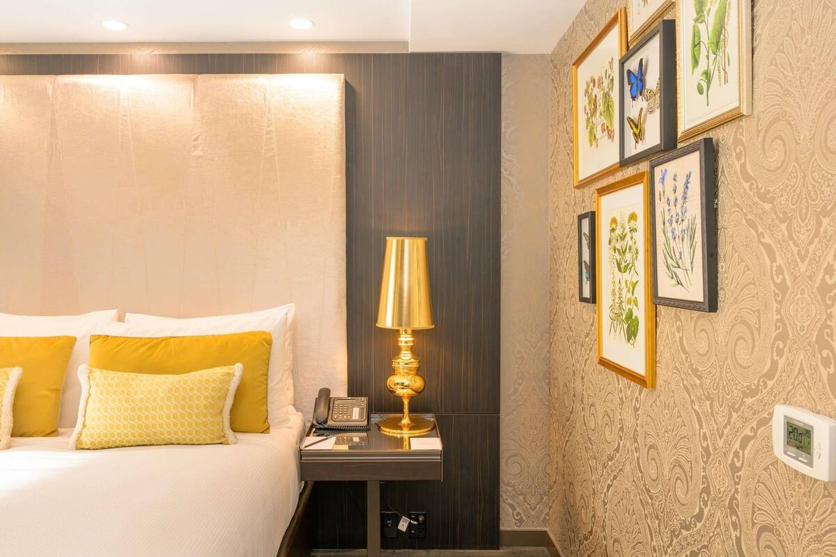 A hotel room with a bed, bedside table with a lamp, phone, and notepad. There are framed botanical prints on the wall.