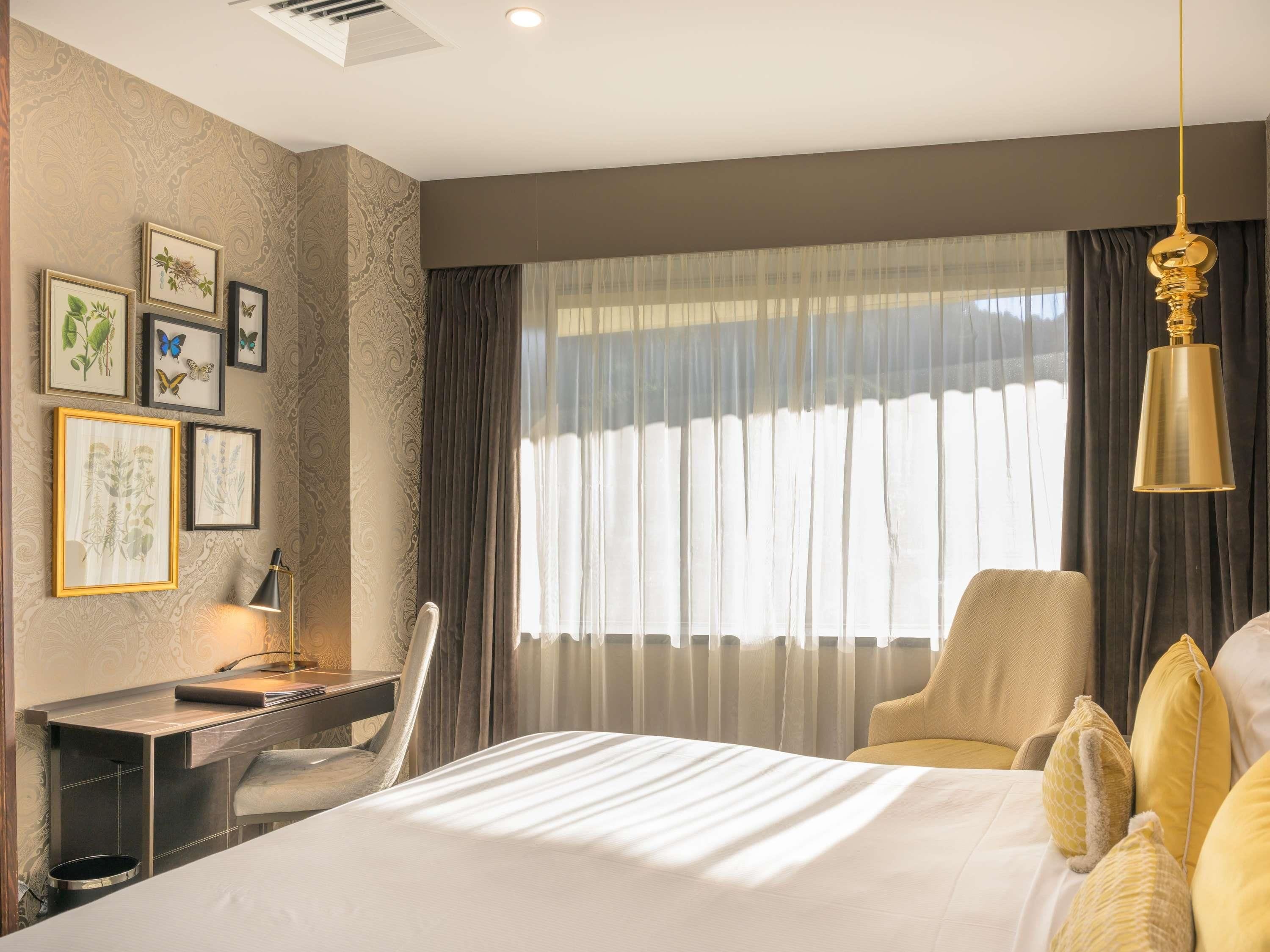 Habitación de hotel con cama, escritorio, silla, lámpara de escritorio y un gran ventanal con cortinas.