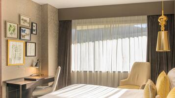 Habitación de hotel con cama, escritorio, silla, lámpara de escritorio y un gran ventanal con cortinas.