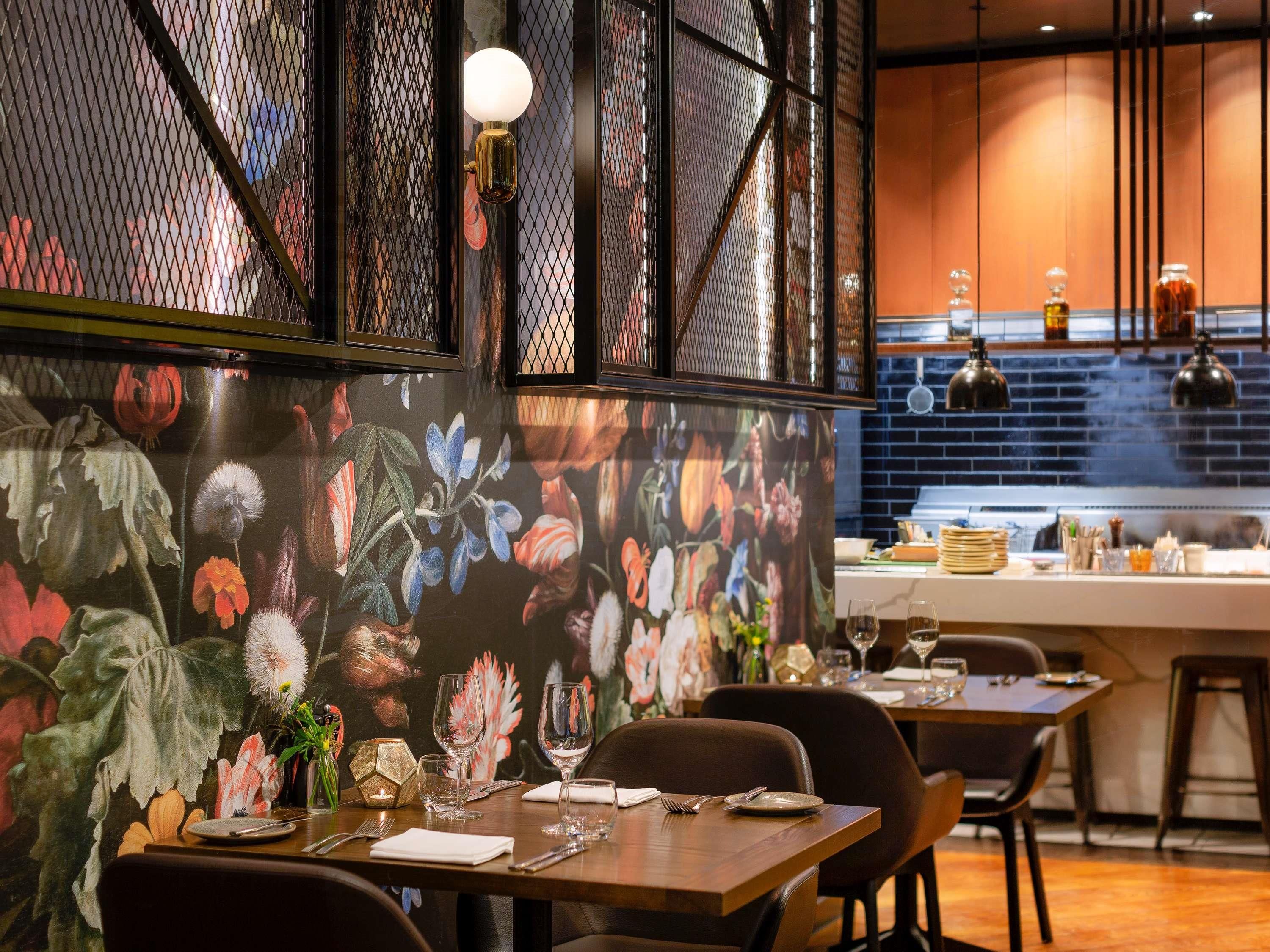 Interior de restaurant amb una taula per a dos, una paret decorativa amb obra floral i una zona de cuina al fons.