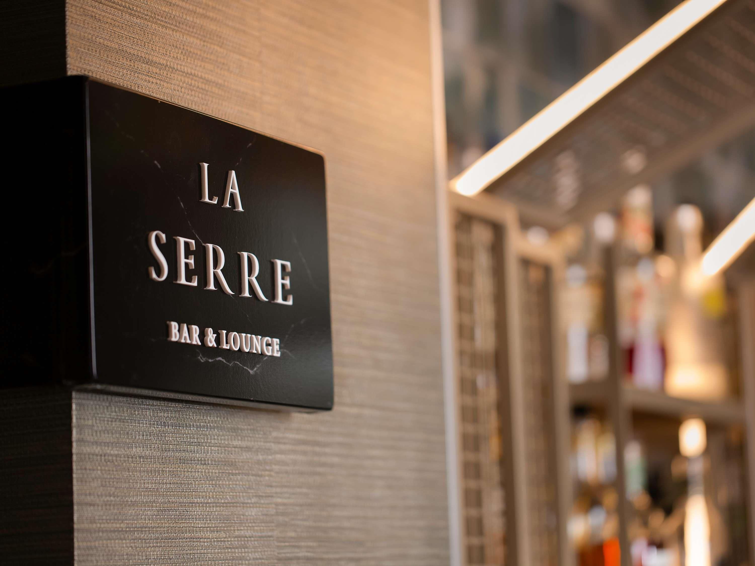 Une enseigne indiquant « LA SERRE BAR & LOUNGE », avec un arrière-plan flou composé d’étagères et de bouteilles.
