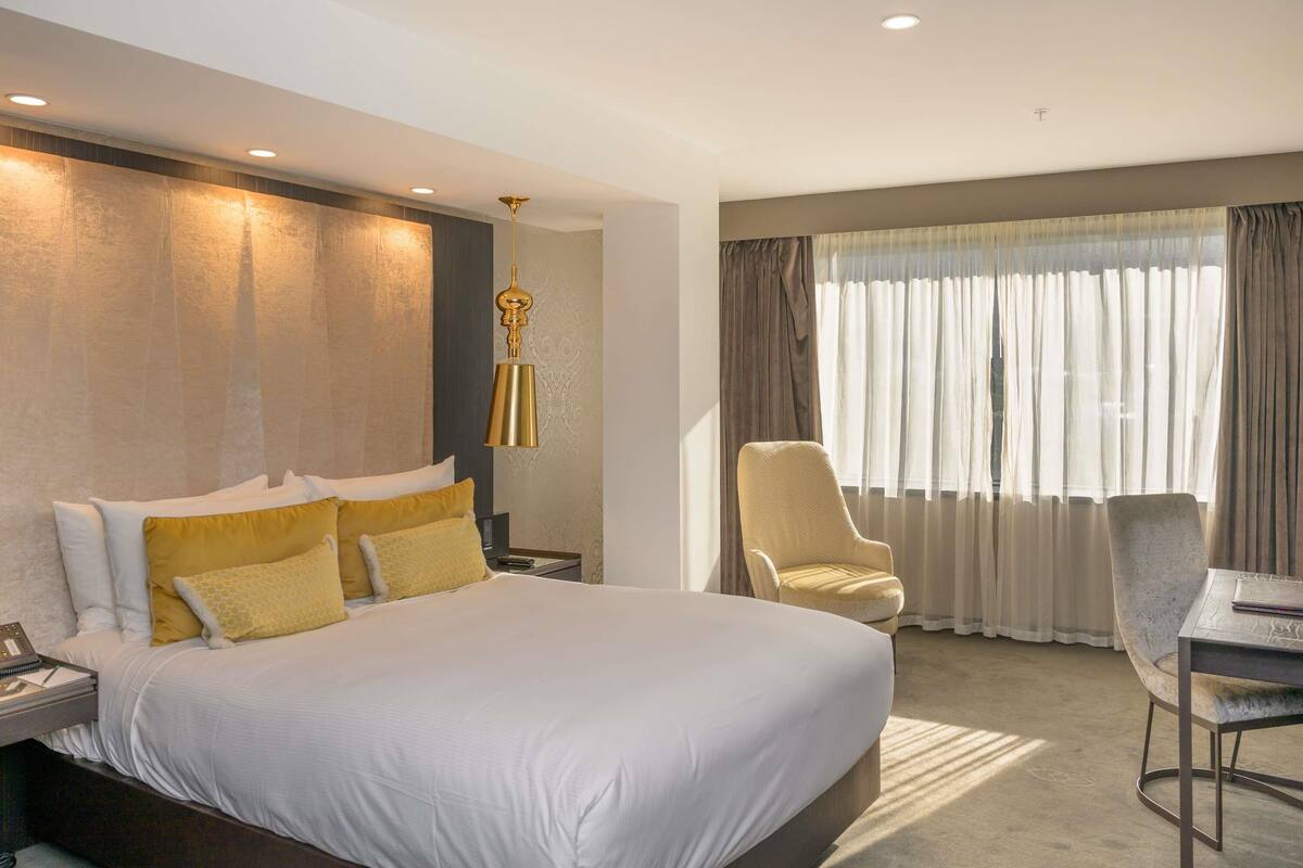 A hotel room with a large bed, a desk with a chair, a desk lamp, and a window with curtains.