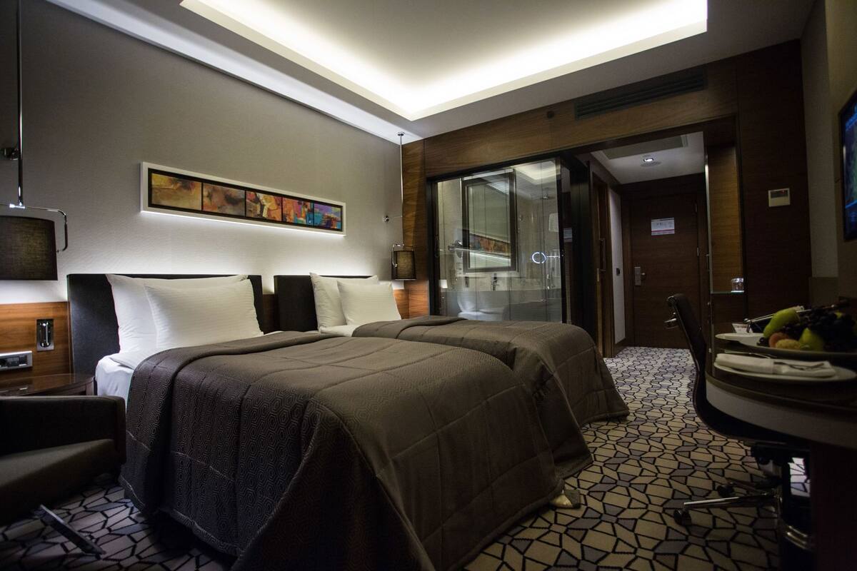 A hotel room with a large bed, a desk, a chair, a small table with fruit, and a bathroom visible through a glass door.