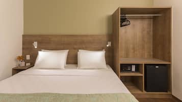 Quarto de hotel com cabeceira de madeira, duas almofadas brancas, cama com colcha estampada, mesa de cabeceira com um vaso de flores, um refrigerador preto pequeno e um guarda-roupa de madeira com cabideiro.