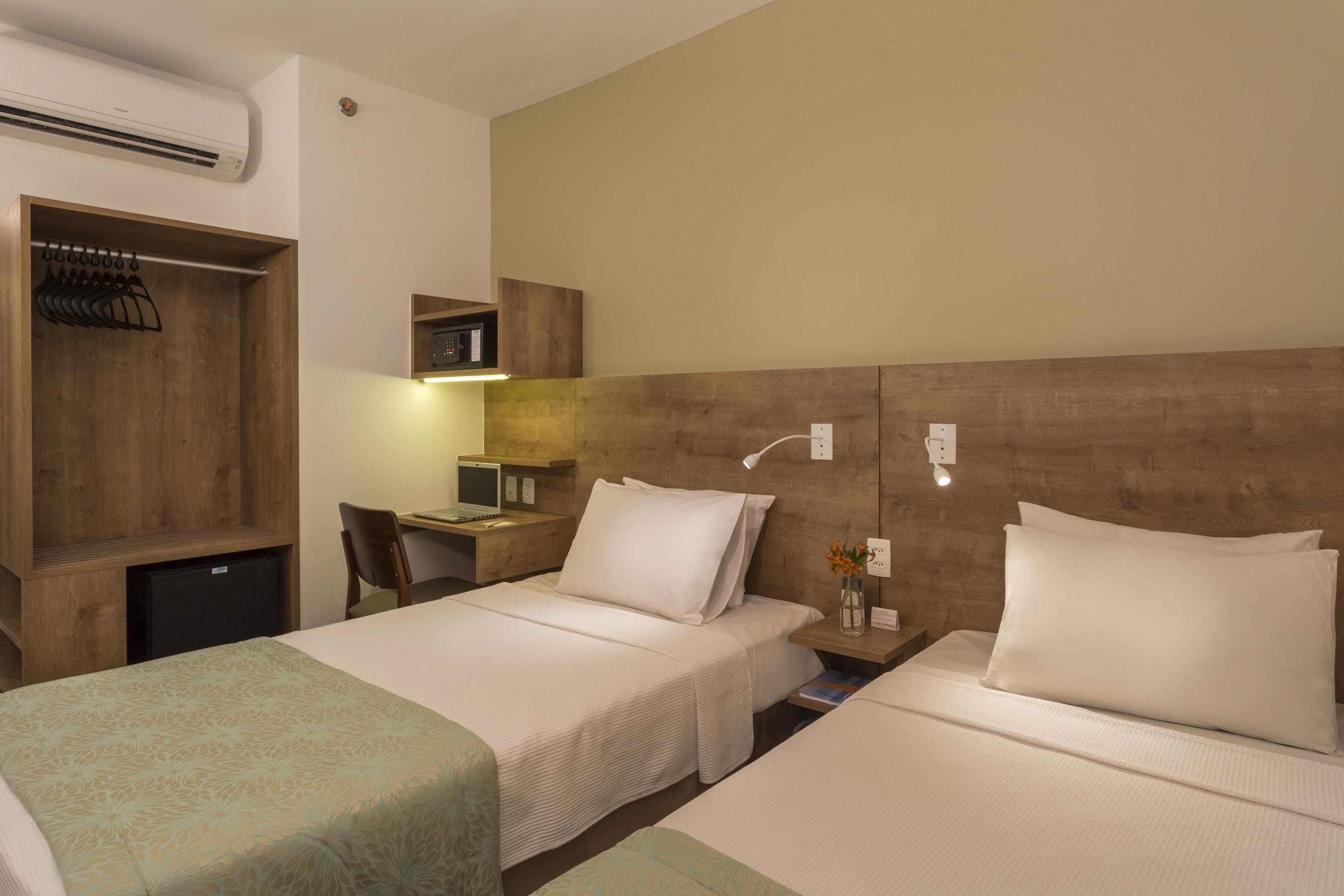 Quarto de hotel com duas camas, uma escrivaninha, uma cadeira, um guarda-roupa e uma pequena luminária de mesa.
