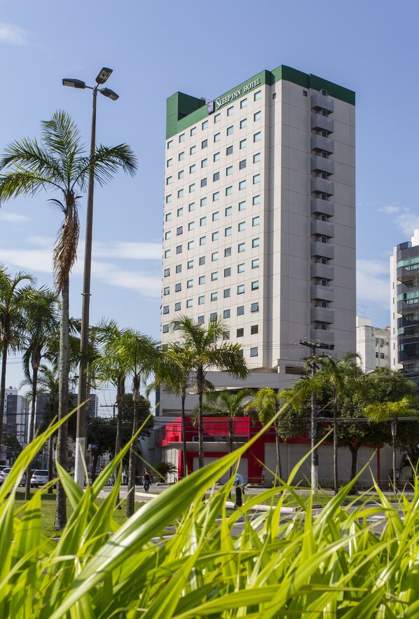 A tall hotel building with multiple floors and balconies, surrounded by palm trees and a clear sky.