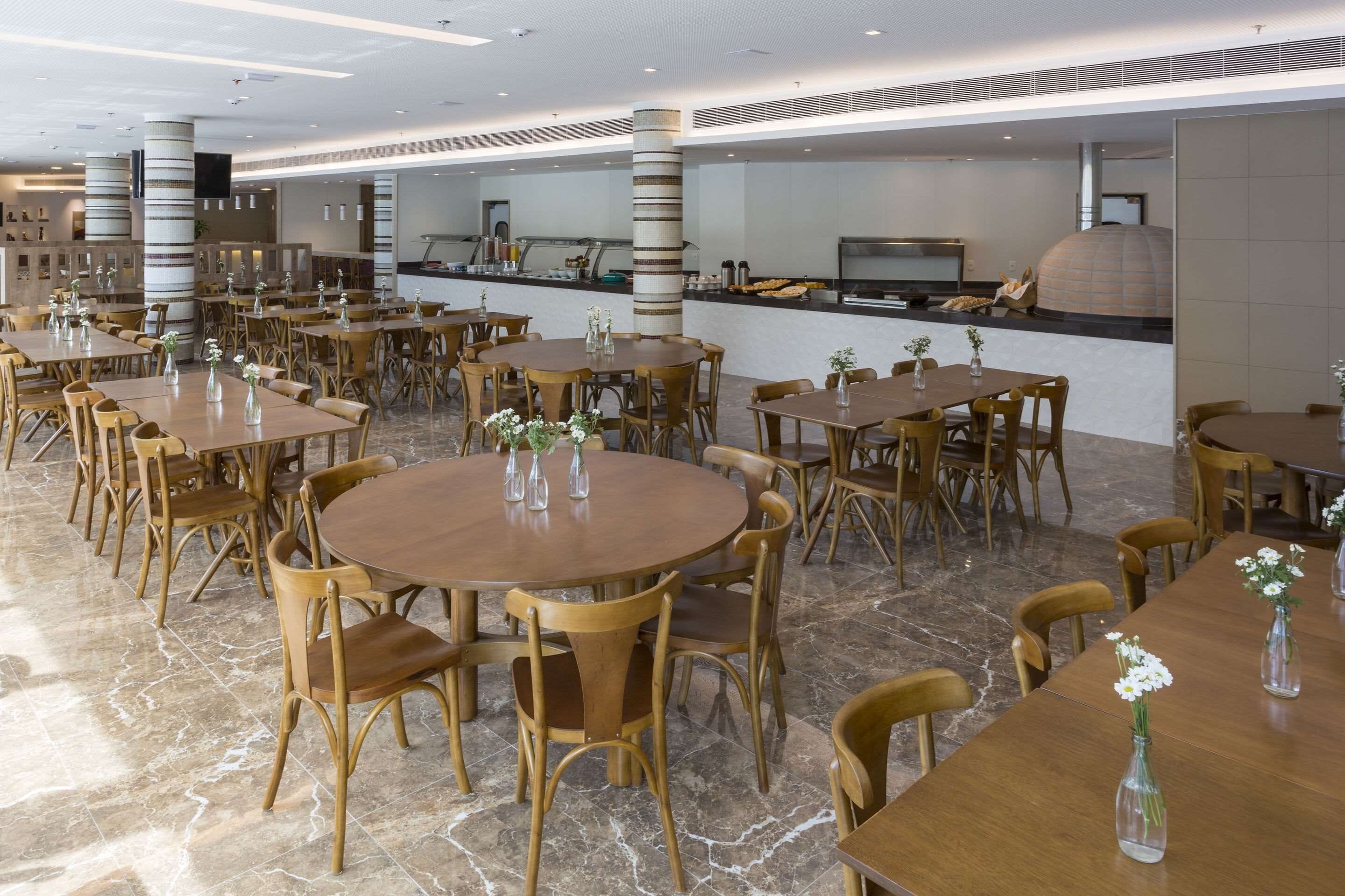 Uma sala de jantar espaçosa com mesas redondas e cadeiras de madeira, uma cozinha ao fundo e um design de teto moderno.