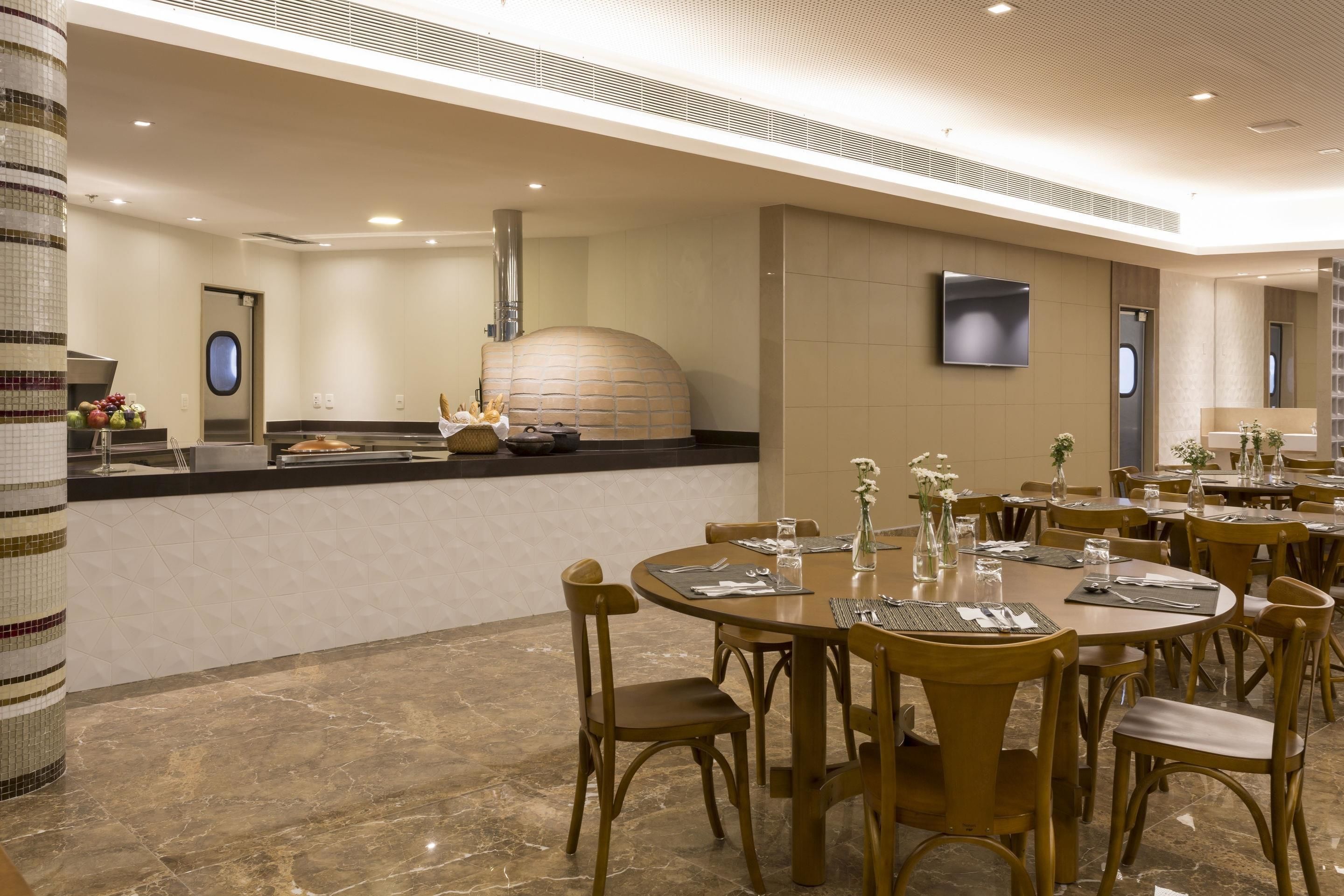 A modern restaurant with round tables, wooden chairs, a central kitchen, and a tiled floor.