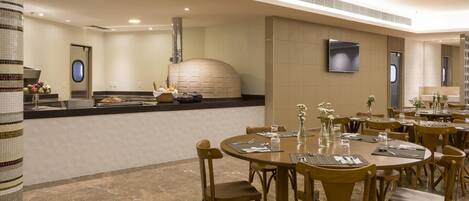A modern restaurant with round tables, wooden chairs, a central kitchen, and a tiled floor.