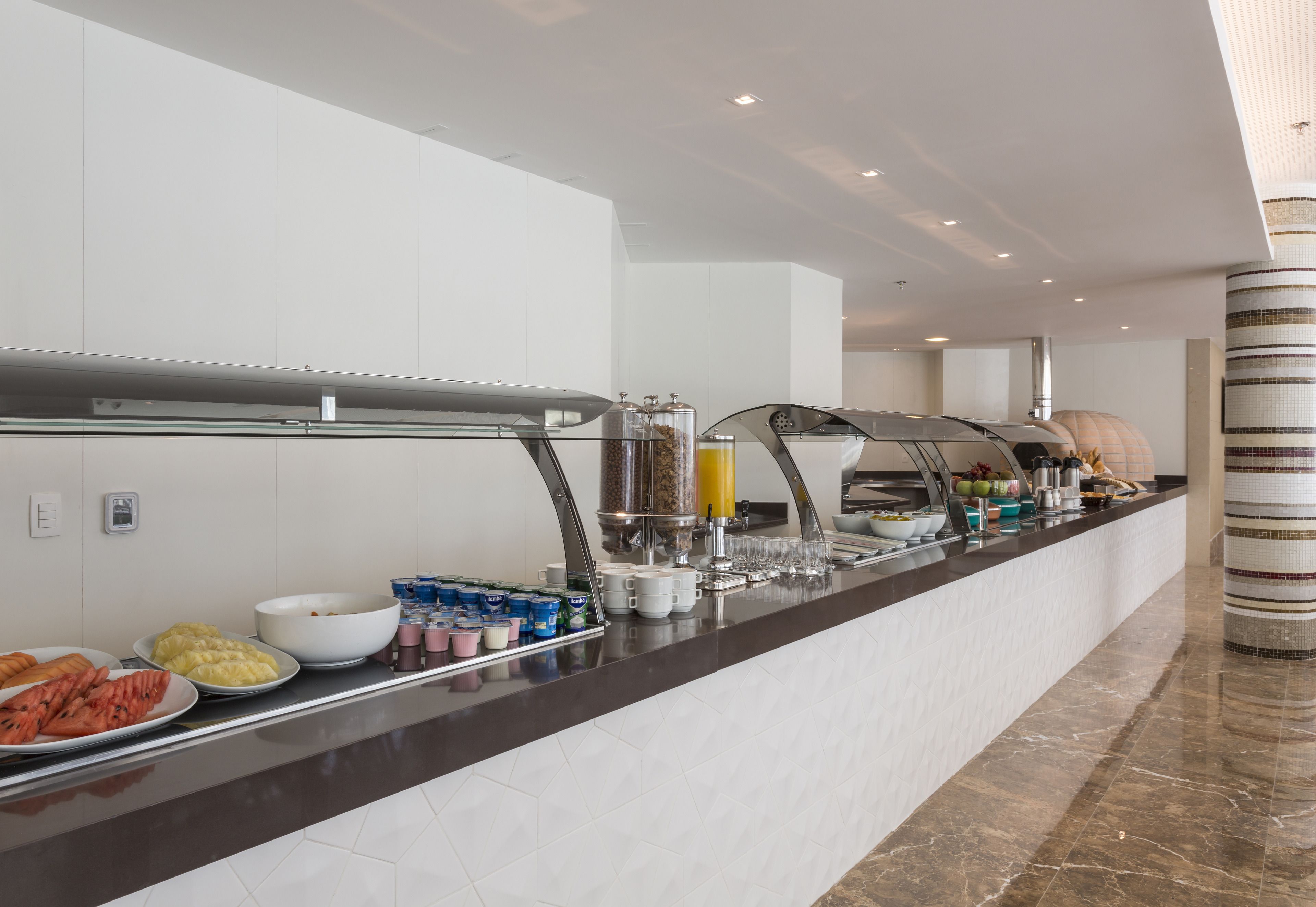 Un moderno mostrador de desayuno de hotel con una variedad de alimentos y bebidas, incluyendo frutas, pasteles y refrescos.