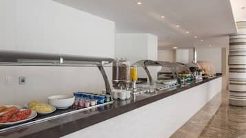 Un moderno mostrador de desayuno de hotel con una variedad de alimentos y bebidas, incluyendo frutas, pasteles y refrescos.