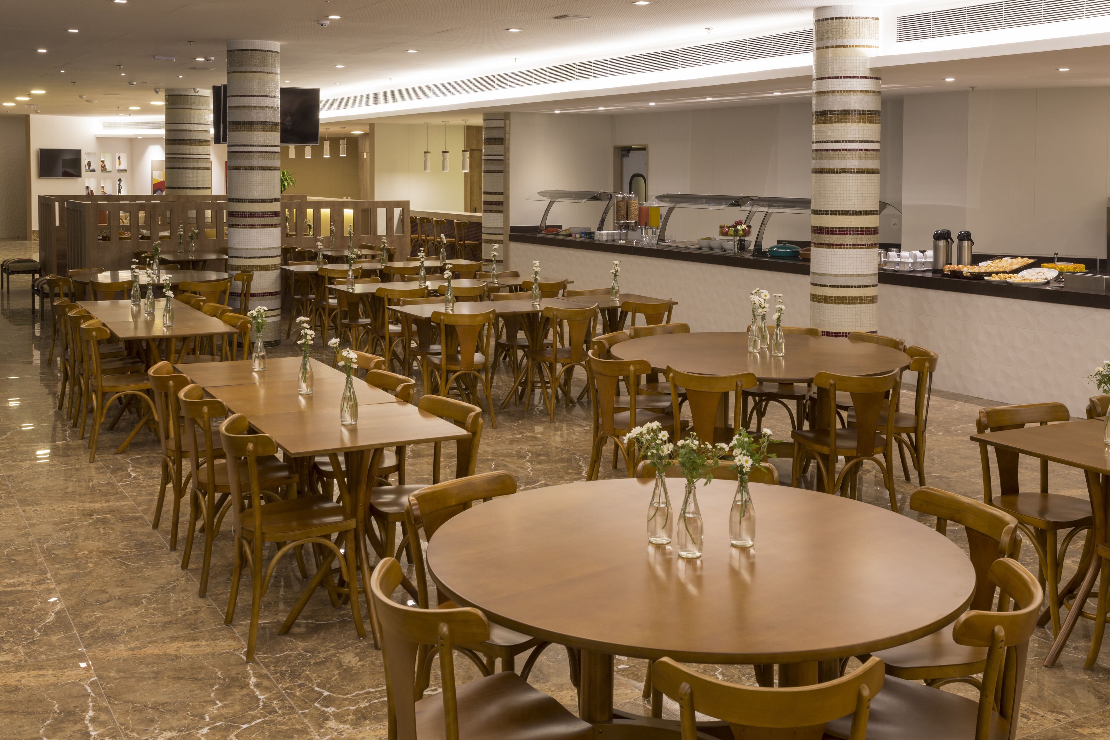 Un amplio comedor con varias mesas y sillas redondas, una barra de buffet y pilares decorativos.