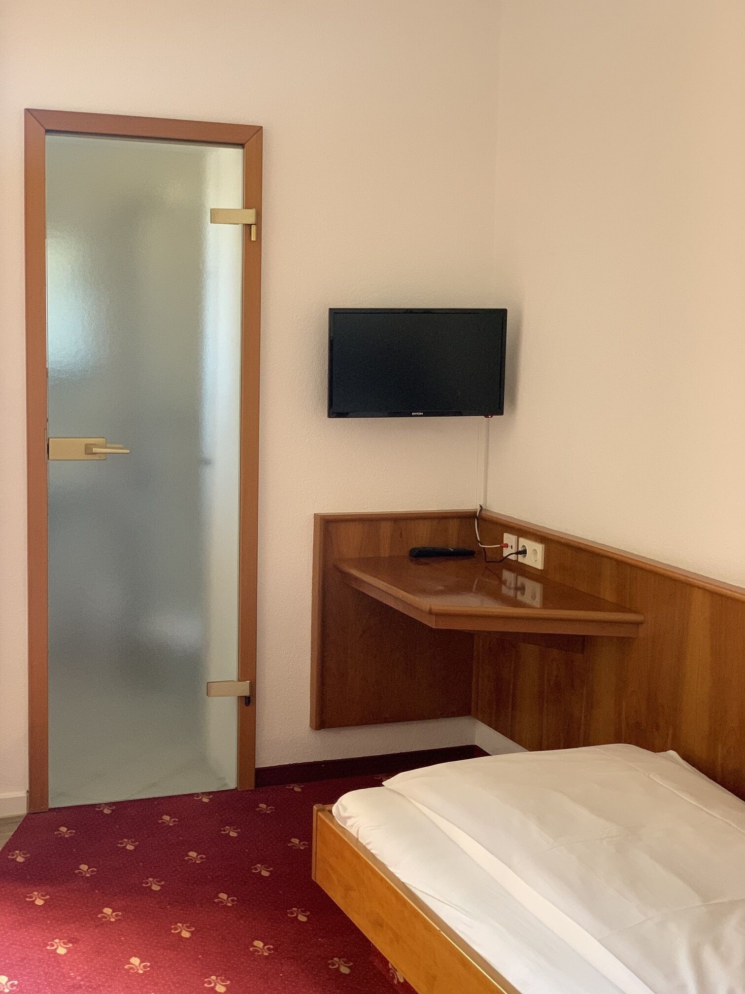Une chambre d’hôtel avec un lit, un téléviseur fixé au mur et une étagère en bois en dessous.