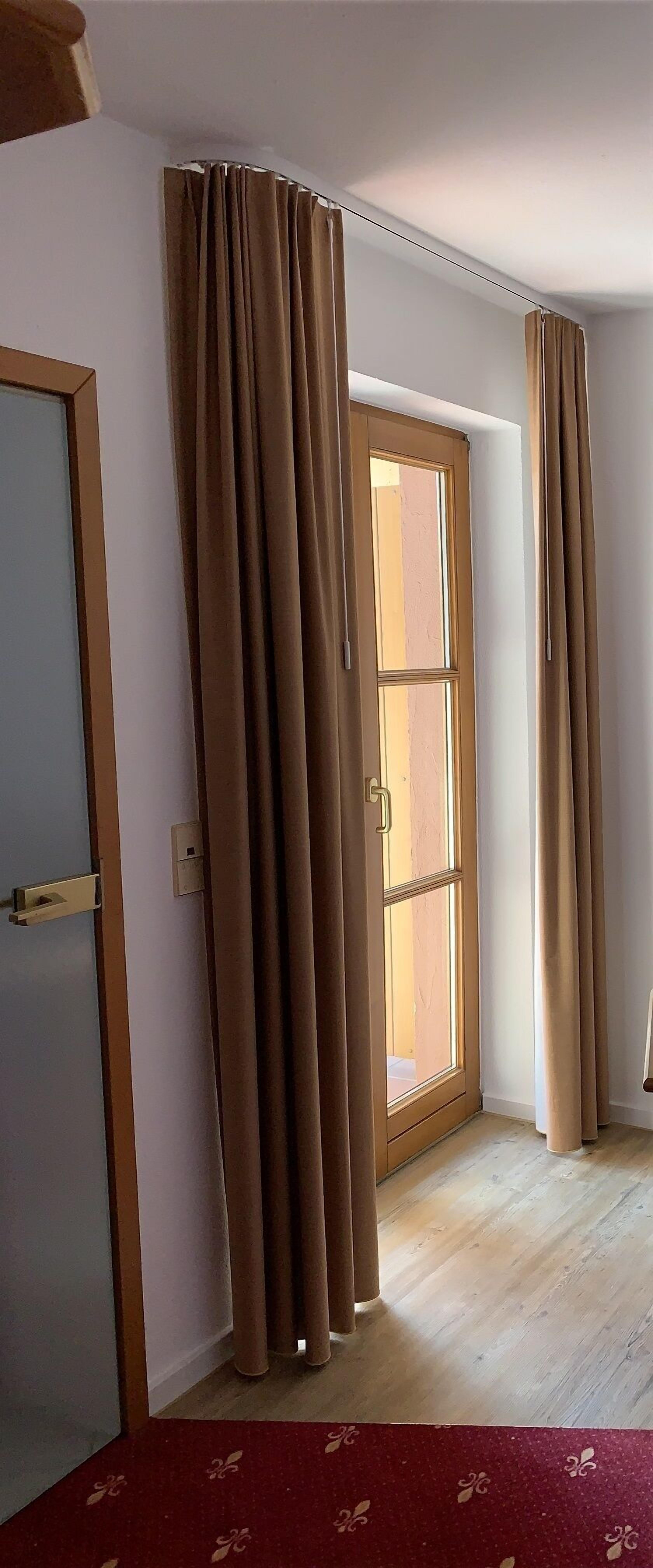 Una habitación con puerta de madera, ventana con cortinas y alfombra roja estampada.