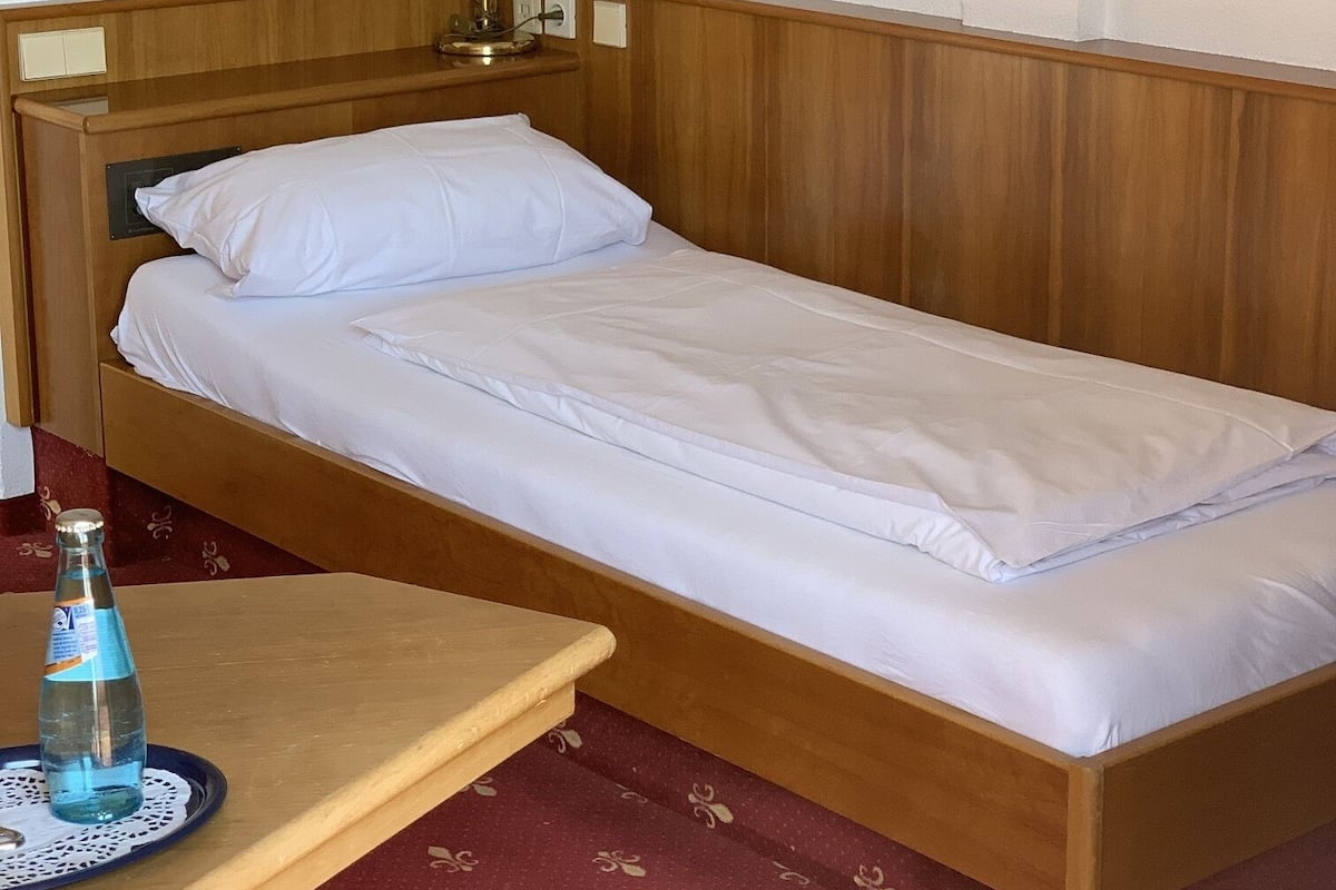 A single bed with a wooden headboard, a bedside table with a bottle and glass, and a wall-mounted lamp.