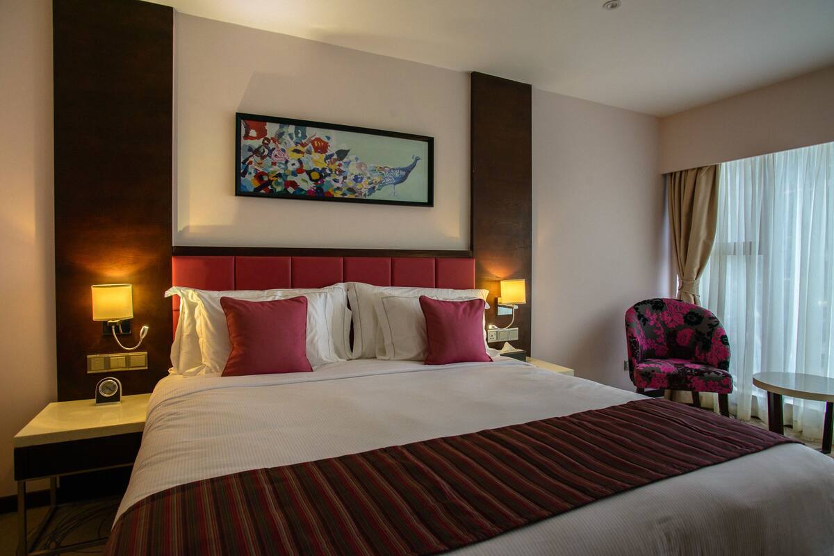 Una habitación de hotel con una cama grande, dos mesitas de noche con lámparas, una silla y un cuadro en la pared.