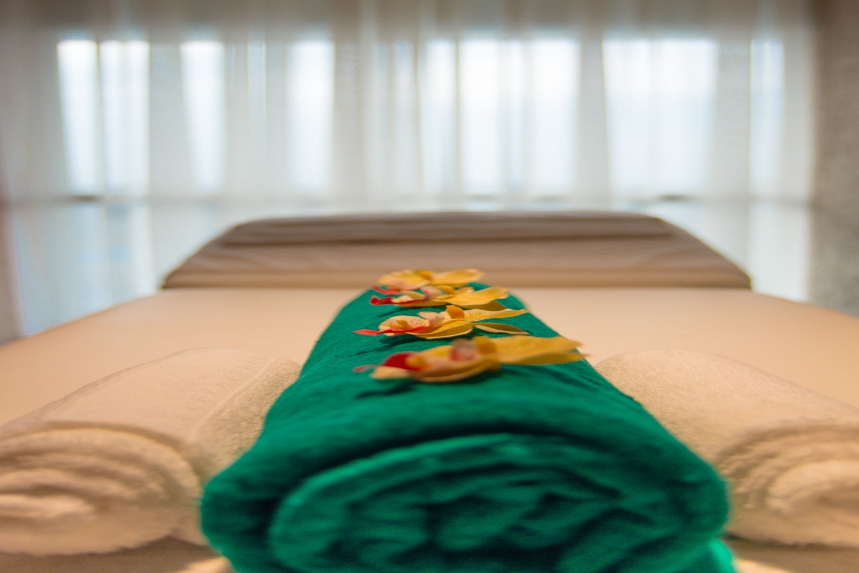 A neatly made bed with a green towel and decorative pillows, a window with blinds in the background.