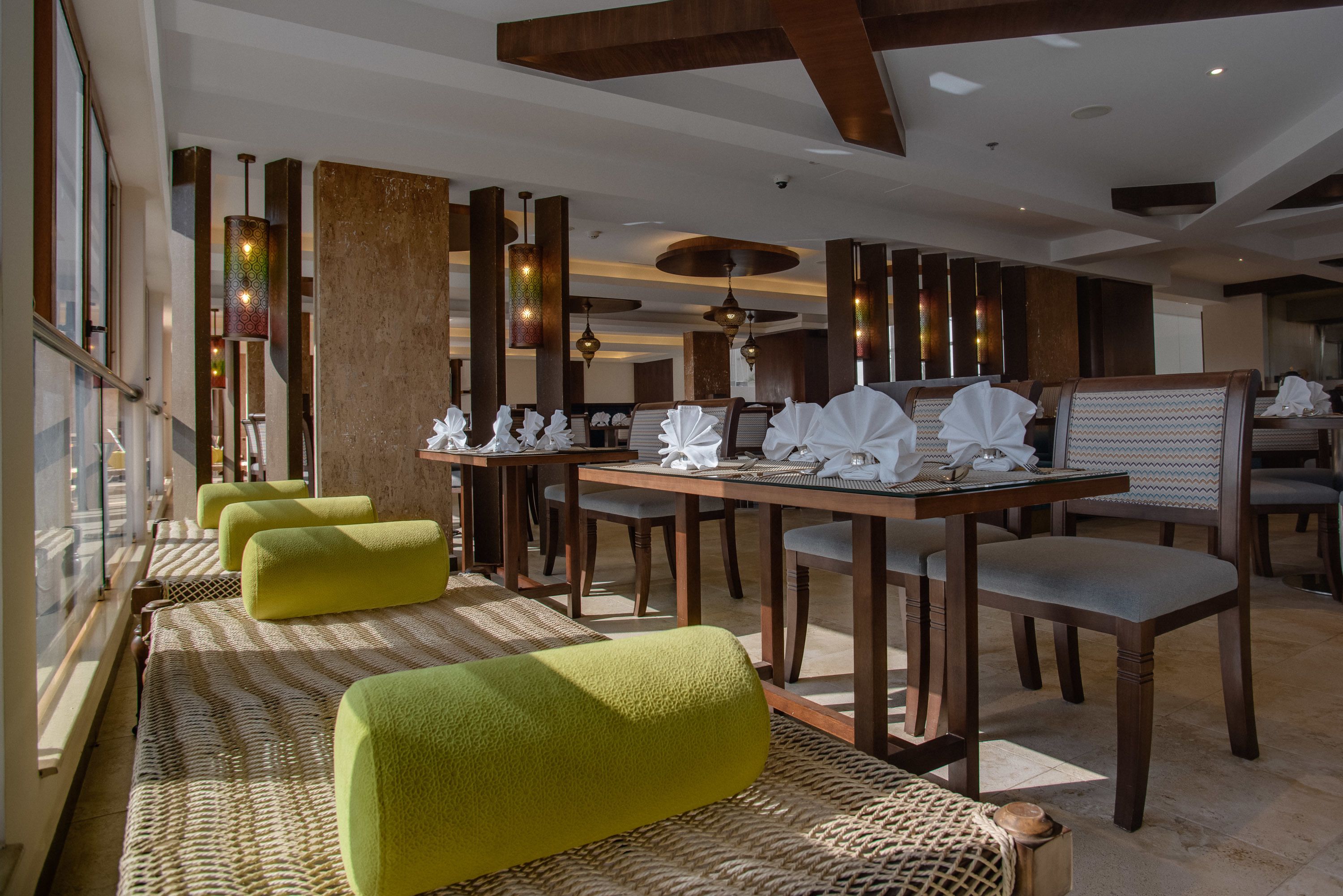 Un restaurante moderno con mesas y sillas de madera, asientos acolchados verdes y luminarias colgantes decorativas.