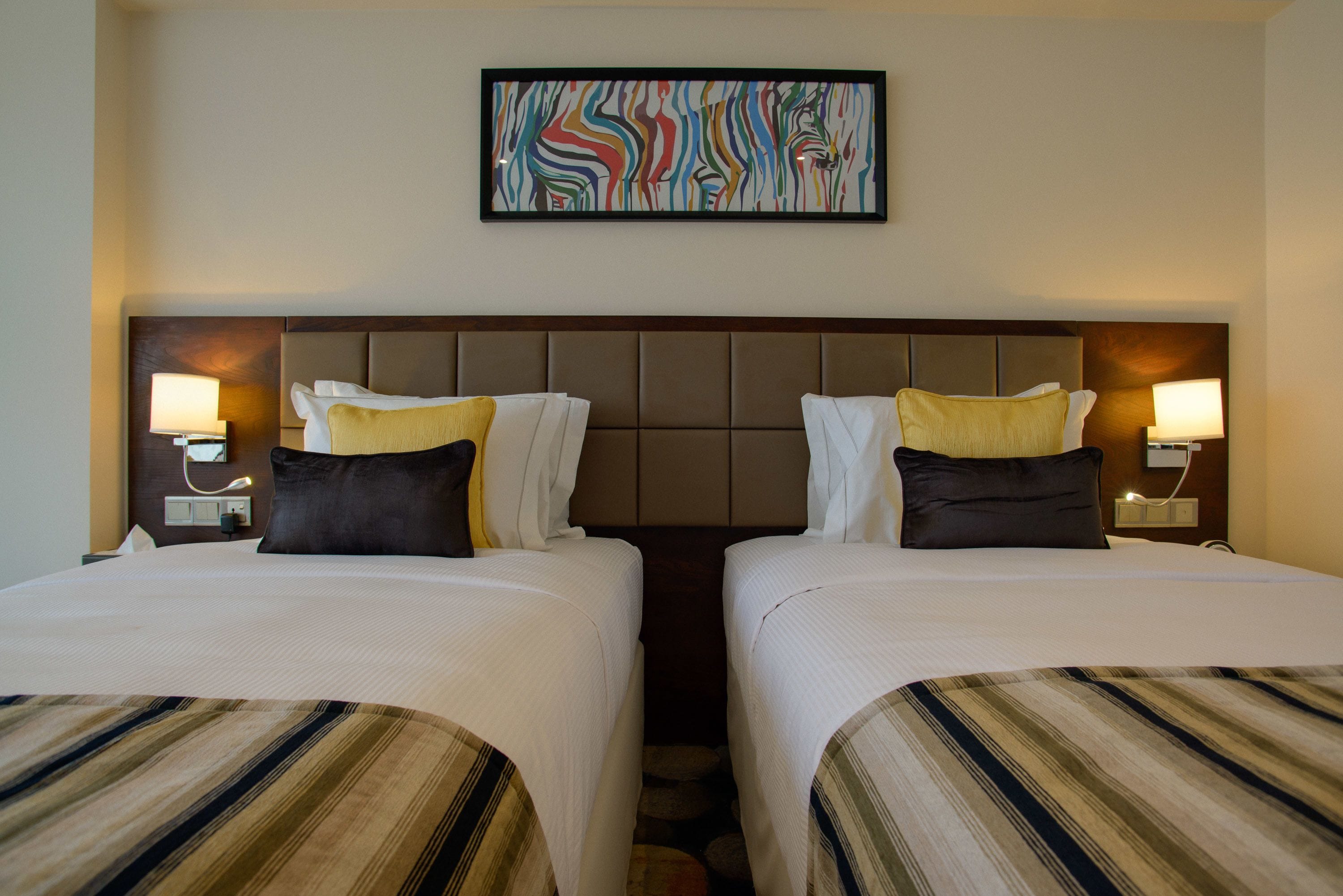 A hotel room with two beds, a headboard, bedside lamps, and a colorful abstract painting on the wall.