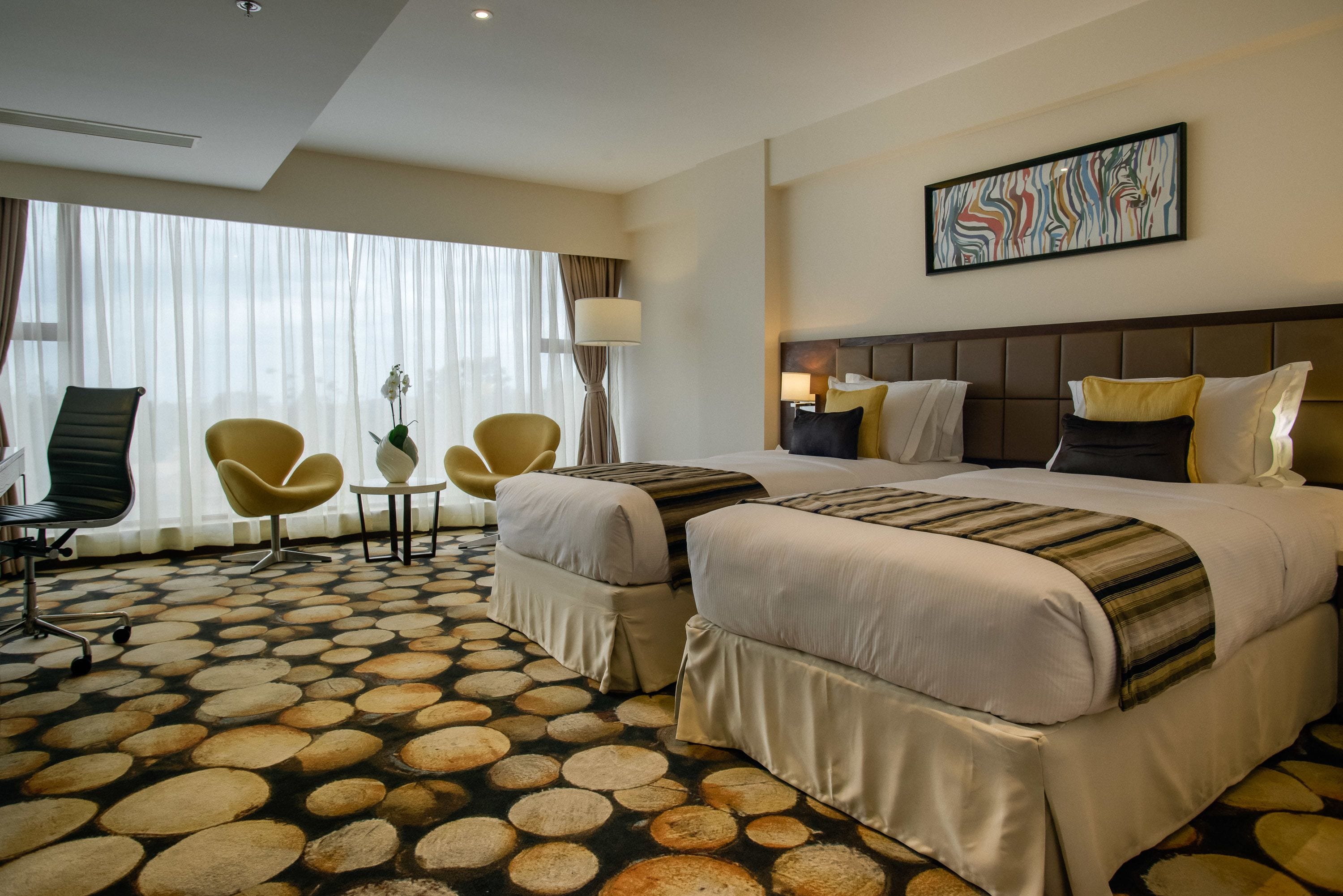 Ein Hotelzimmer mit zwei Betten, einem großen Fenster mit transparenten Vorhängen, einem modernen Sessel, einem kleinen Tisch und einem dekorativen Bodenmuster.