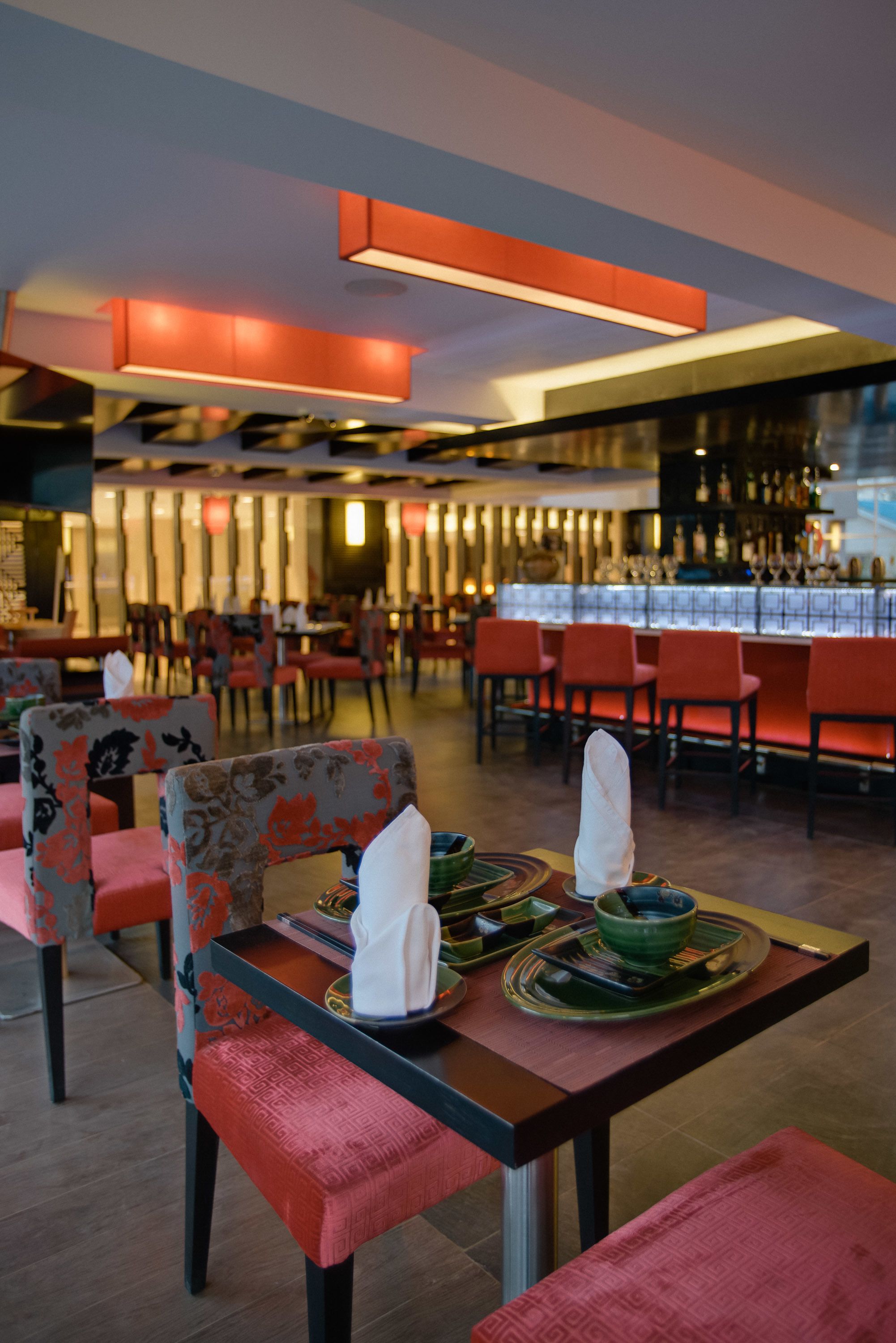 A restaurant with red upholstered chairs, a bar area in the background, and a table set with plates and bowls.