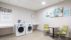 A guest laundry area with two washing machines, a small table, and two chairs. - Holiday Inn Express and Suites Lincoln I - 80 by IHG (Lincoln)