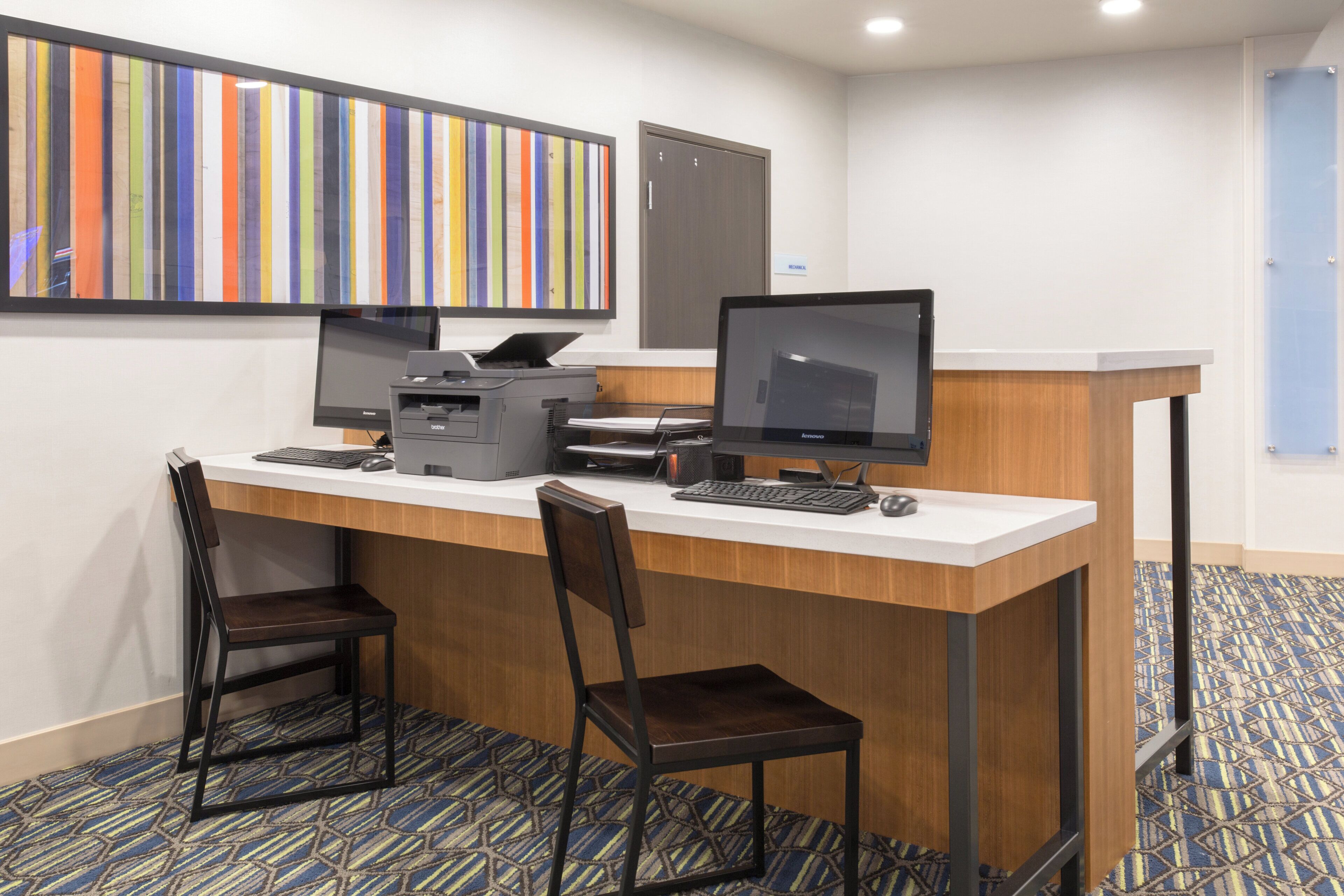Een hotel receptiebalie met twee computerschermen, een printer en een toetsenbord. Er staan twee stoelen en een klein tafeltje.