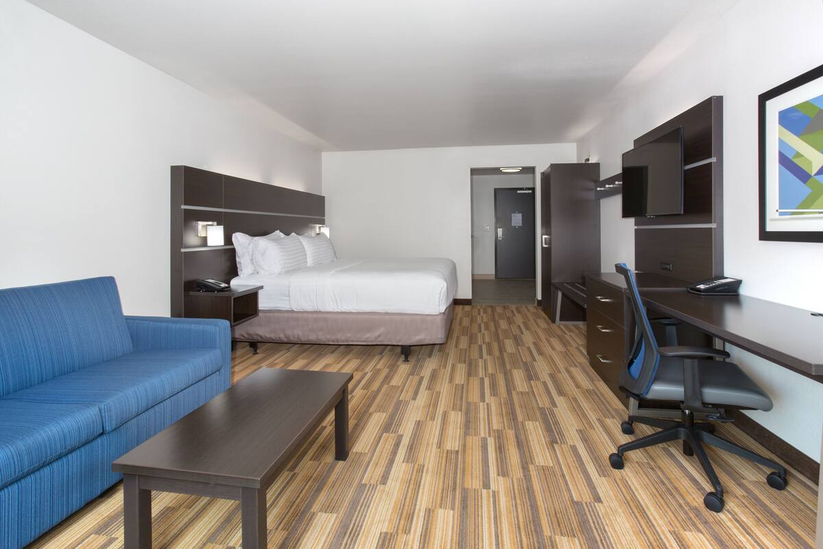 A hotel room with a bed, a blue sofa, a desk with a chair, a TV mounted on the wall, and a wooden floor.