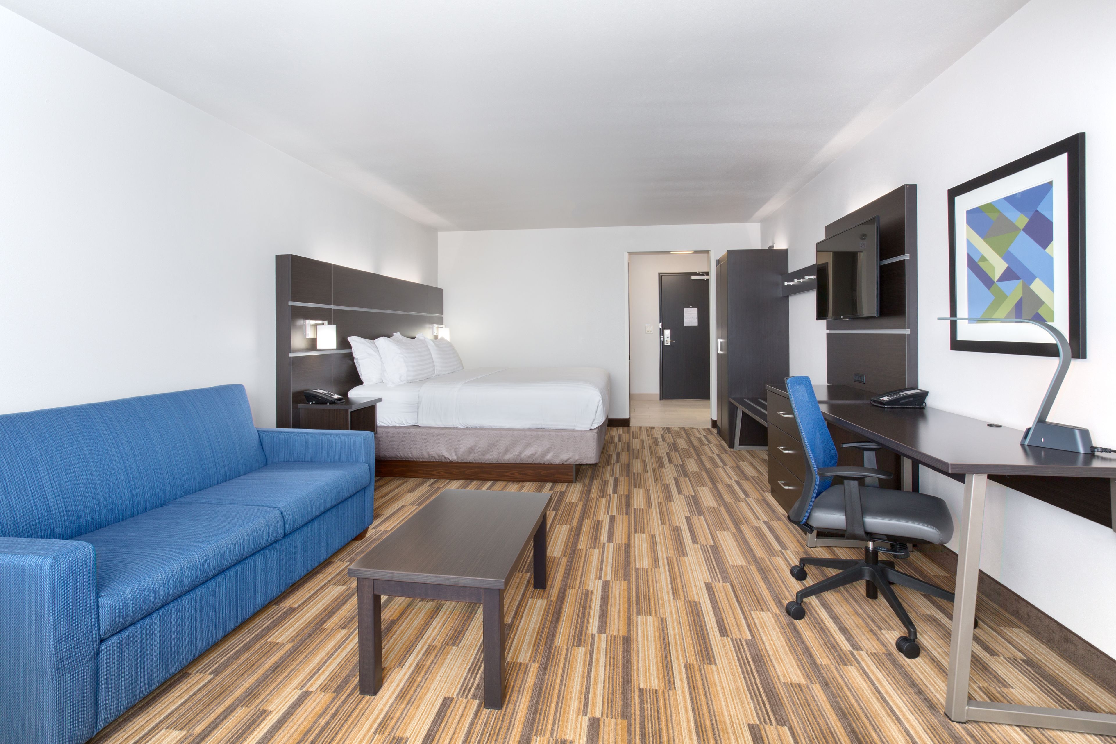 A hotel room with a bed, a blue sofa, a desk with a chair, a TV, and a wooden floor.