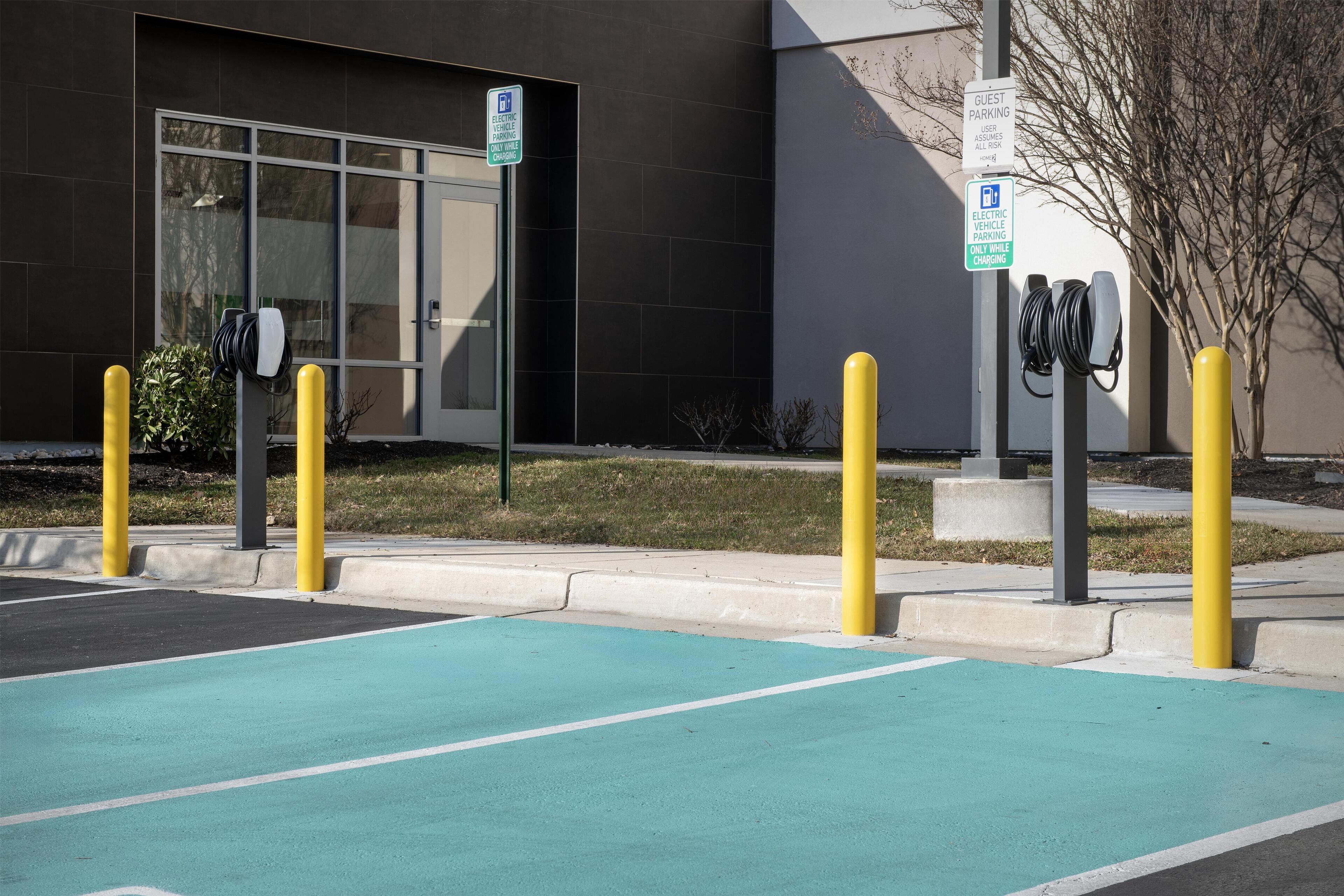Una estación de carga para vehículos eléctricos con un espacio de estacionamiento azul, bolardos amarillos y un edificio con un amplio ventanal y señalización.