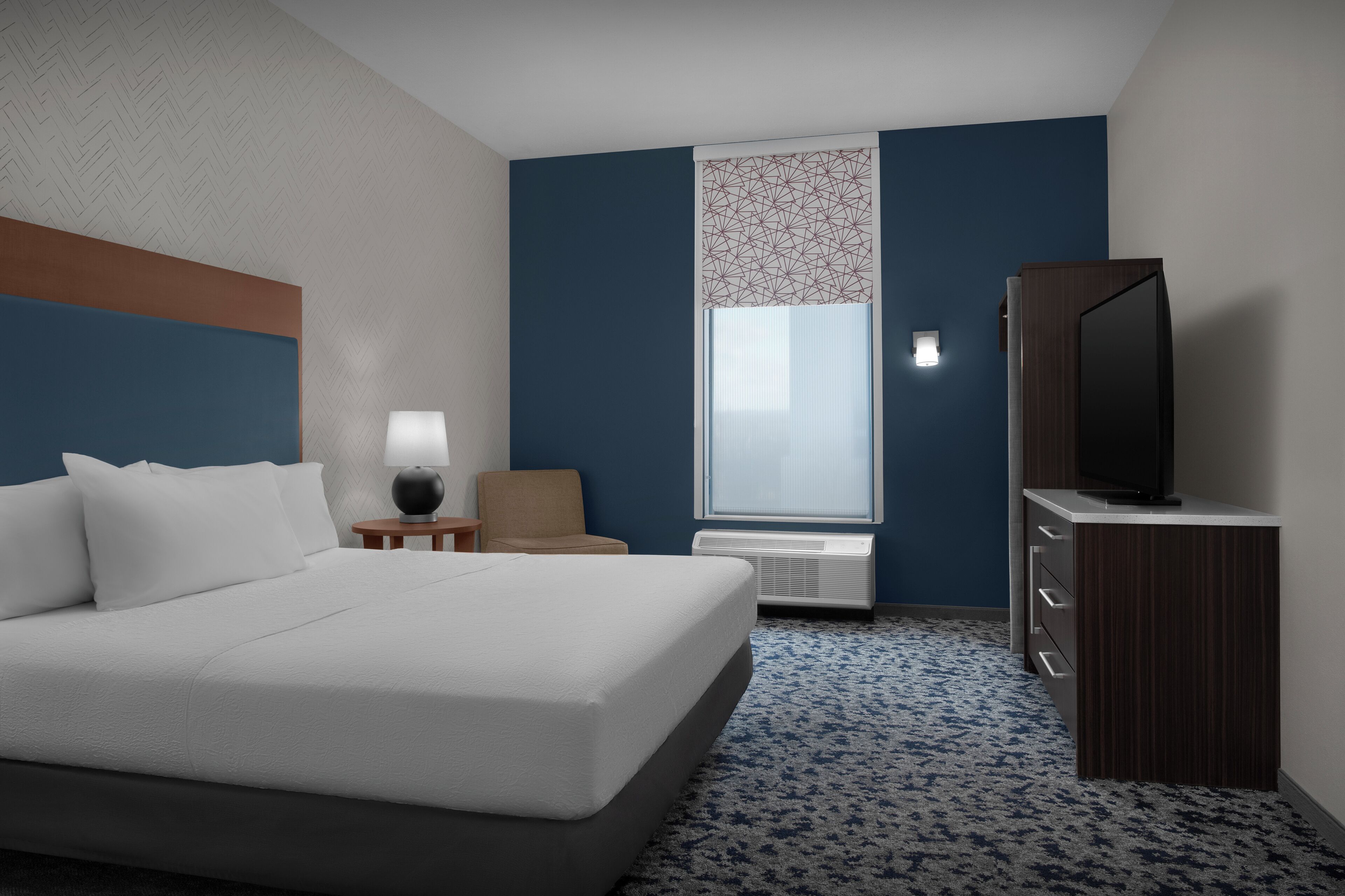 A hotel room with a large bed, a television on a wooden cabinet, a chair, a lamp, and a window with a patterned curtain.