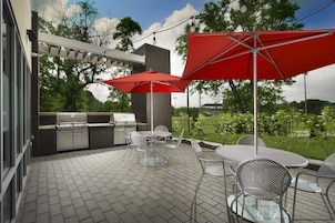 Un patio al aire libre con una parrilla, mesas, sillas y un paraguas rojo.