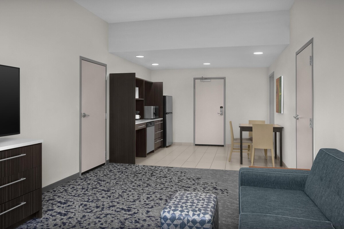 Habitación de hotel moderna con televisión de pantalla plana, sofá, alfombra estampada, kitchenette y zona de comedor.