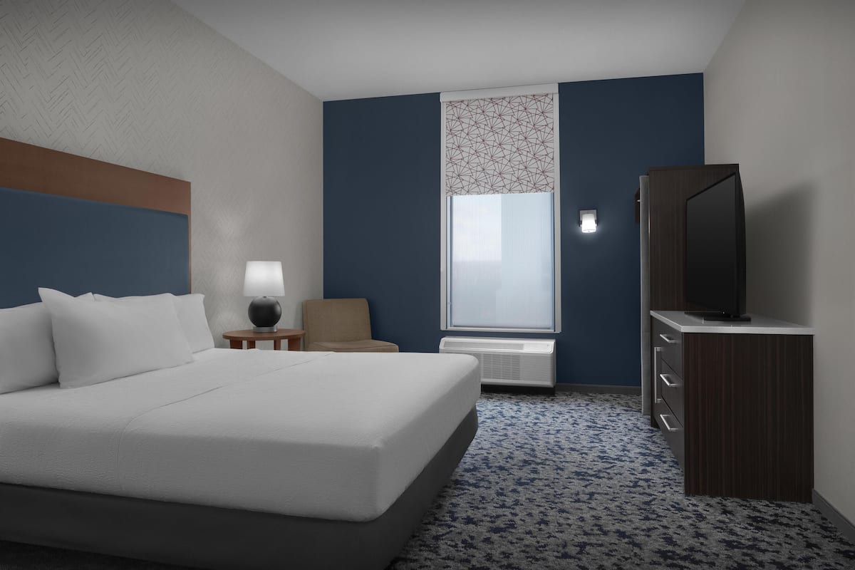 Habitació d'hotel amb un llit, un televisor sobre un moble de fusta, una cadira, una làmpada i una finestra amb un disseny geomètric.