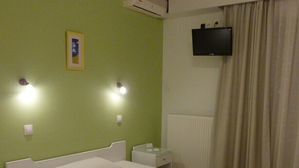 A neatly made bed with white linens, a bedside table with a lamp, and a wall-mounted TV. - Melissanthi Hotel (Nea Propontida)