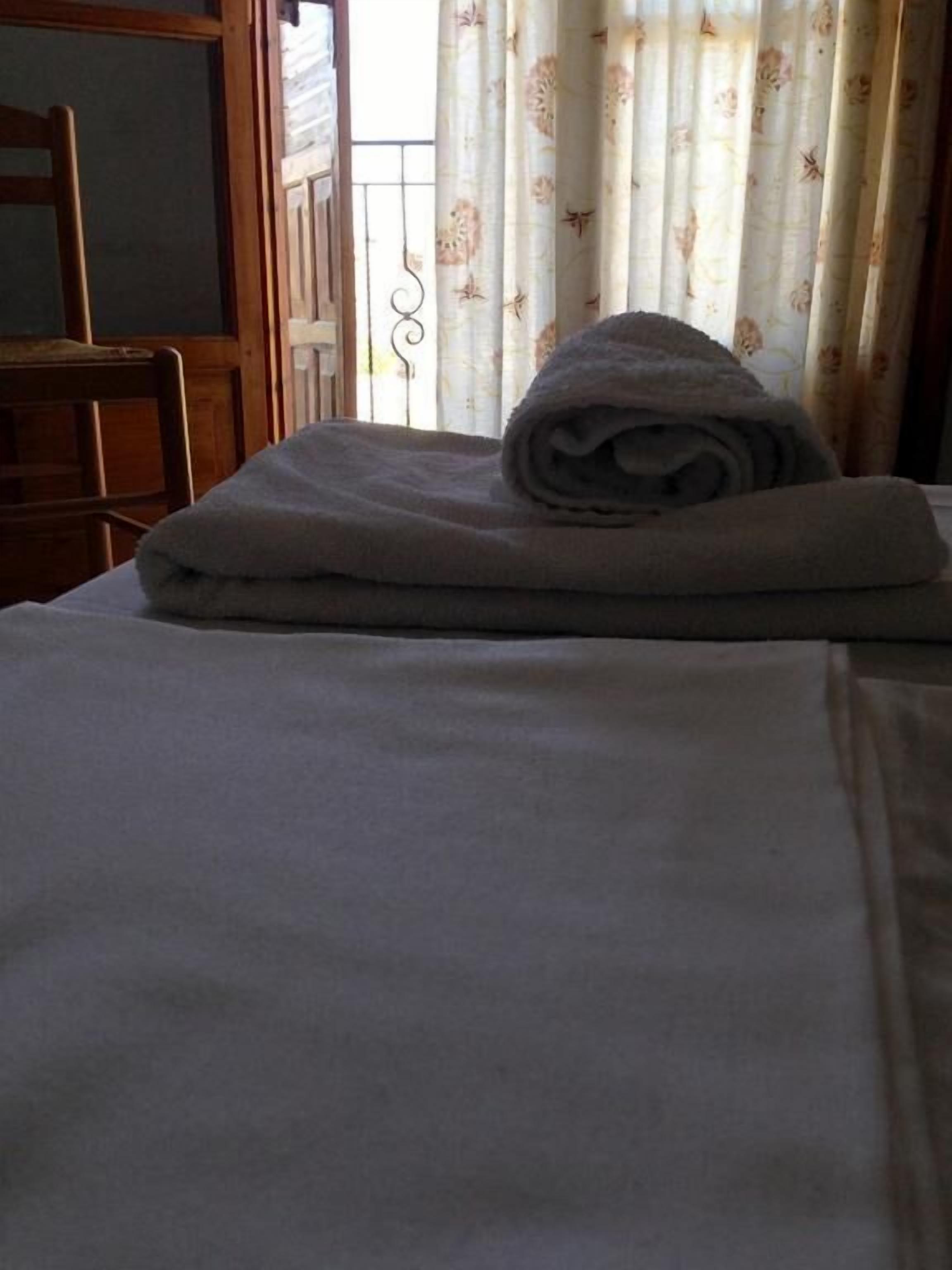 Una cama bien hecha con una toalla doblada encima, una silla de madera y una ventana con cortinas florales.