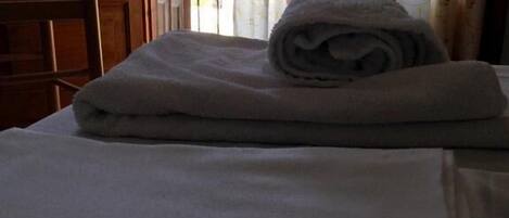 Una cama bien hecha con una toalla doblada encima, una silla de madera y una ventana con cortinas florales.