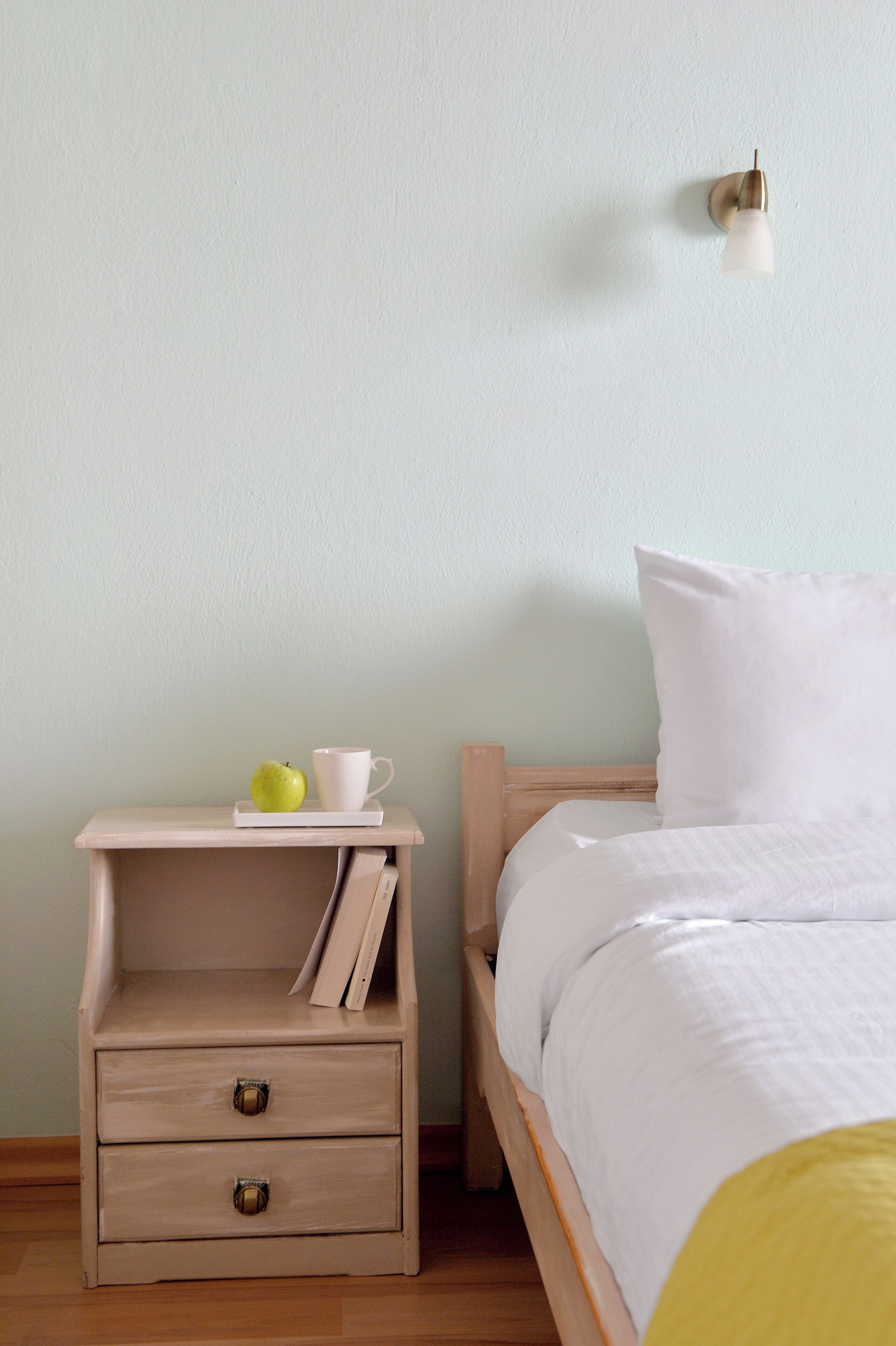A bedroom with a wooden nightstand, a bed with white bedding, a wall-mounted light fixture, and a small shelf with a cup and apple.