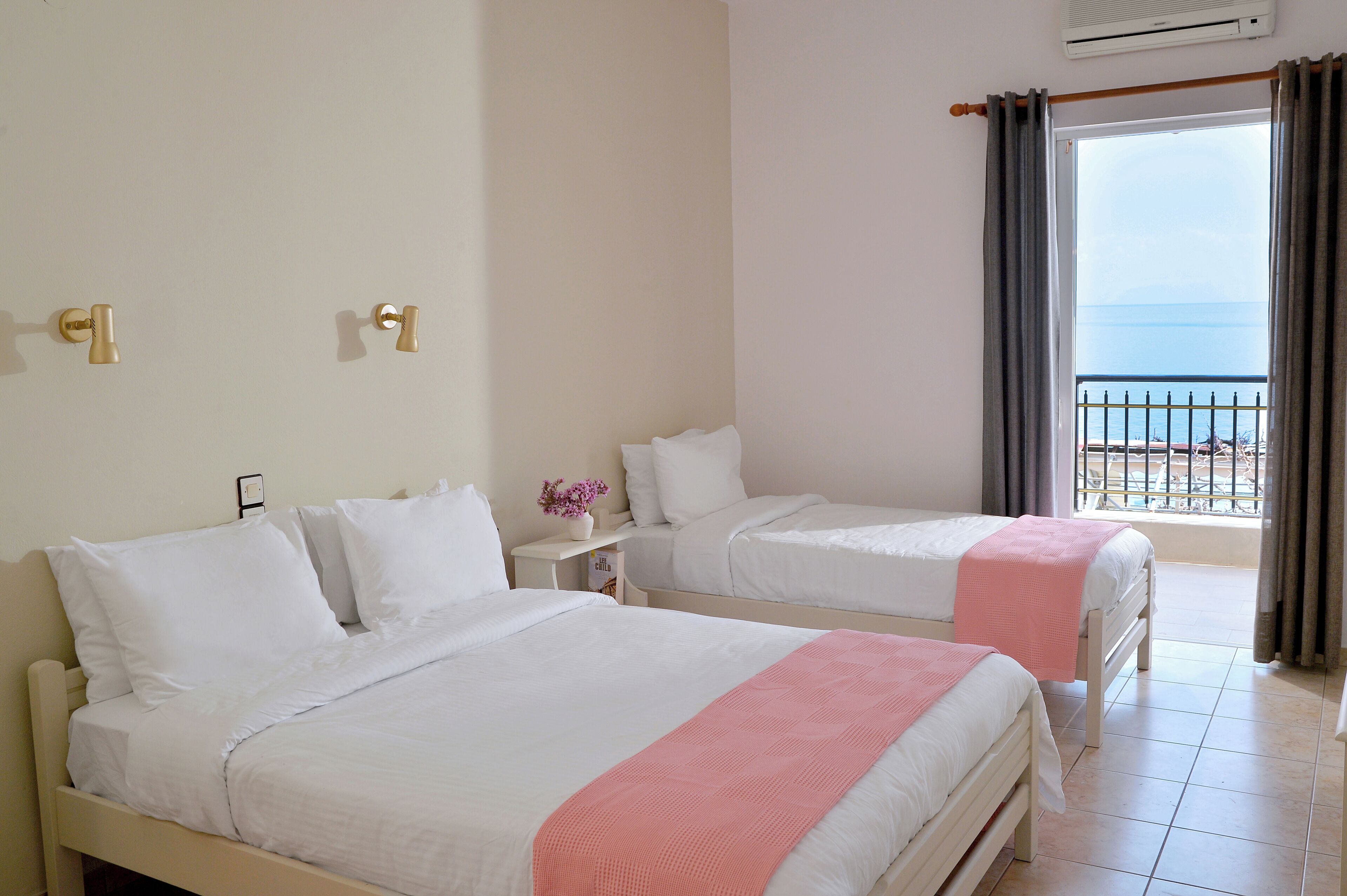 Quarto de hotel com duas camas, uma pequena mesa de cabeceira com um vaso de flores e uma varanda com vista para o mar.
