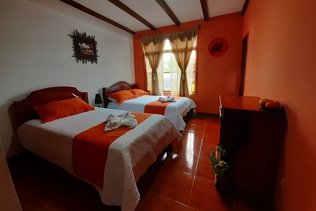 A hotel room with two beds, orange bedding, a wooden headboard, a small desk, and a plant.