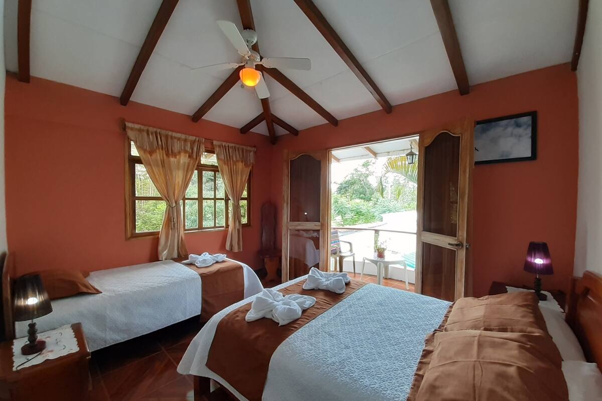 A hotel room with two beds, a ceiling fan, a window with curtains, a small table with a chair, and a door leading outside.